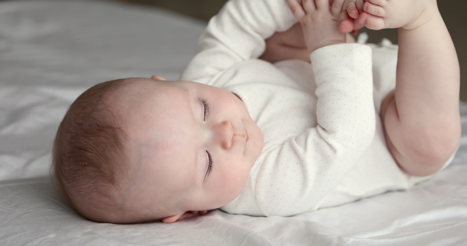 Close up side view cutie three-month-old newborn in white bodysuit lying alone on bed. Adorable baby rests on bedsheets, moving, play with toes. Infancy, healthcare and paediatrics, growth concept Royalty-Free Stock Footage #1078681559