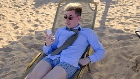 A young funny businessman in a shirt, tie, shorts and sunglasses communicates via video link lying on the beach on the sun lounger using a mobile phone, slow motion, 4k