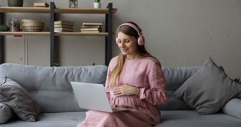 Pregnant woman in headphones sit on couch makes videocall, receive consultation from gynaecologist via videoconference. Online medical counsel at quarantine. Virtual remote meeting with family concept