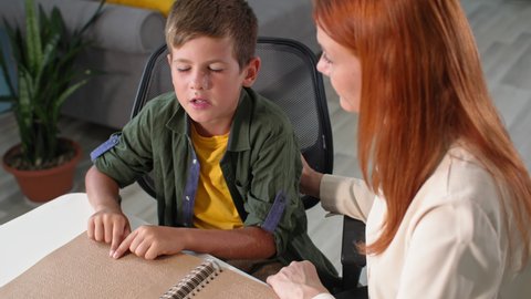 blind child learns to read Braille, woman teacher watches as boy kid moves his fingers over the font