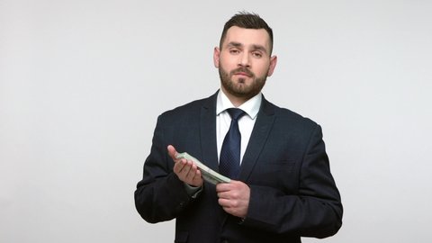 Portrait of satisfied bearded businessman in black official style suit standing with dollars banknotes, looking at camera with proud expression. Indoor studio shot isolated on gray background.