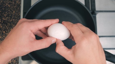 Male hands breaks a chicken egg over a hot frying pan without oil, top view