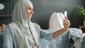Beautiful hijabi girl in scarf is chatting and waving hand during video call from apartment room. Muslim people and modern technology concept.