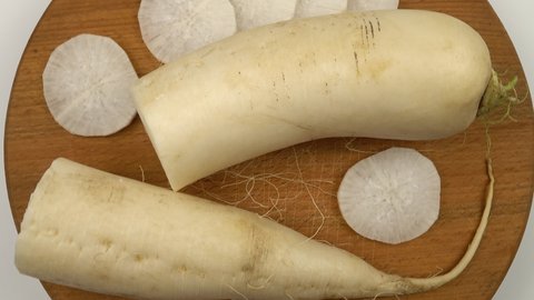 Halves of uncleaned daikon radish and thin slices of radish lie on a wooden cutting board. Fresh raw daikon radishes, popular ingredients in vegetable salads and snacks