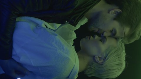 Portrait of two passionate lovers hugging, kissing in trendy colorful neon illumination indoors close up. Happy feelings, sensual embrace, romantic desire of young loving couple. Vertical video format
