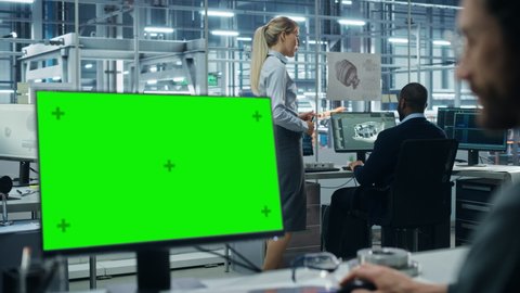 Car Factory Office: Male Automotive Engineer Sitting at His Desk Working on Green Screen Chroma Key Computer. Automated Robot Arm Assembly Line Manufacturing. Over Shoulder Close-up Arc Shot