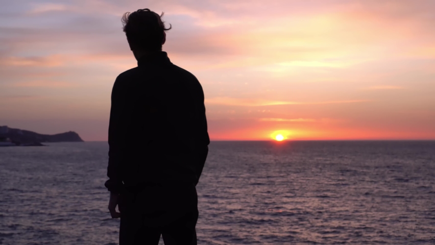 Sad young alone man looks thoughtfully at the ocean. Beautiful sunrise background. Travel and self-isolation, social distancing. Nature, sea, beach, morning Royalty-Free Stock Footage #1078717436