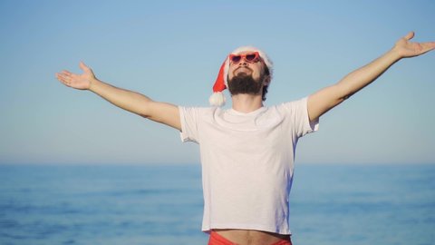 Happy man in red Santa Claus hat raises his hands to the sun by the sea. Funny man with a beard wearing red glasses. concept of New Year's holiday in a warm country on the beach