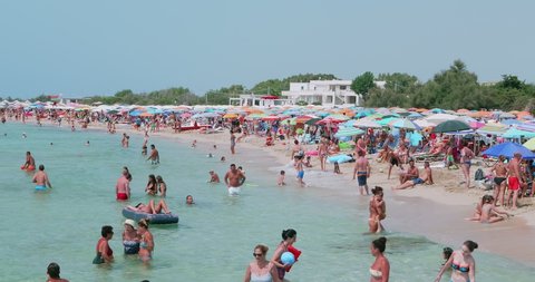 Italy, August 2021: tourists and bathers crowded and gathered on a beach of Marina di Pulsano, near Taranto. Lots of people in the sea and lots of colorful umbrellas. Puglia. - Italy