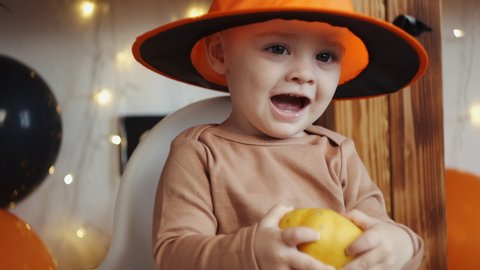 Halloween home family party concept. Portrait of adorable baby wearing Halloween hat holding little pumpkin and smiling. Happy caucasian little kid celebrating Halloween at home.