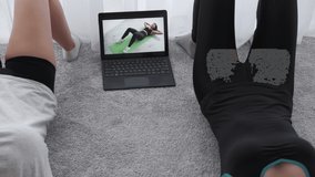 Online sport. Home training. Video lesson. Fitness blog. Unrecognizable two female friends doing abs workout with trainer on laptop screen in light room interior.