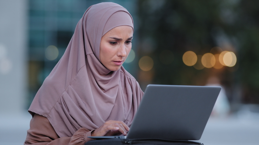 Excited young muslim woman winner looks at laptop celebrates online success sits outdoors in city. Euphoric islamic lady islam girl in hijab gets new distance job opportunity reads good news victory Royalty-Free Stock Footage #1078730525