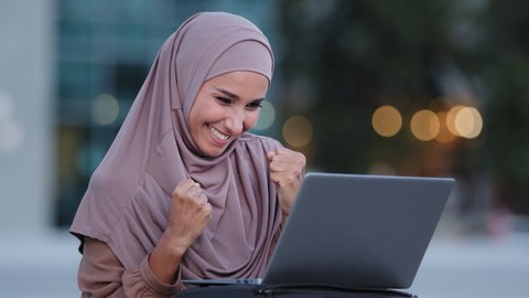Excited young muslim woman winner looks at laptop celebrates online success sits outdoors in city. Euphoric islamic lady islam girl in hijab gets new distance job opportunity reads good news victory
