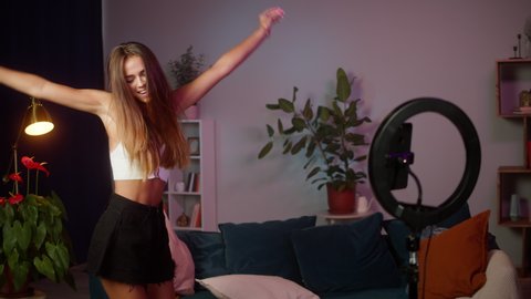 Young woman recording dance video on modern smartphone camera with professional light in living room. Female student or teenager dancing for musical clip. Blogger making music content for channel.