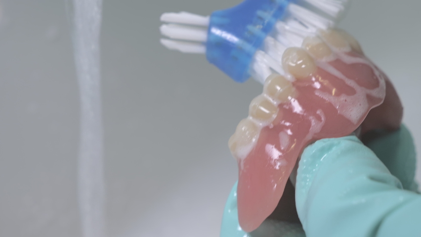 Closeup shot of cleaning dentures using a brush. Running water in the background and rubber gloves being used for better hygiene. | Shutterstock HD Video #1078735445