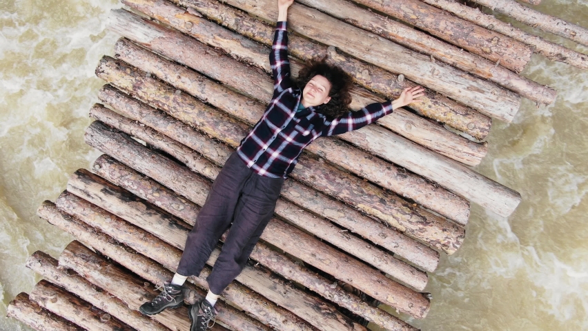 Aerial view of a female tourist in a plaid shirt lying on an old wooden bridge over a stormy mountain river, the drone takes off and rotates
