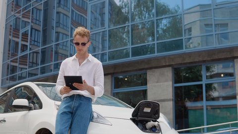 Stylish modern young curly man stands near an electric car charging and uses his tablet