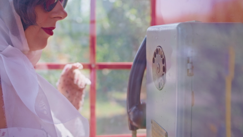 beautiful young woman happily talking on the phone in a red telephone booth in English style. girl in a white dress and glasses. pin up. dials a phone number, close-up.