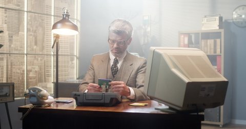 Retro caucasian male working in vintage interior of office. Man in glasses sitting at old computer and examining diskettes at the table. Serious businessman of 80's