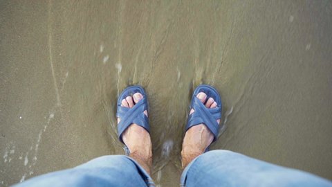First-person view of feet in flip-flops. The sea waves arrive and wash over your feet. The topic of tourism and travel.