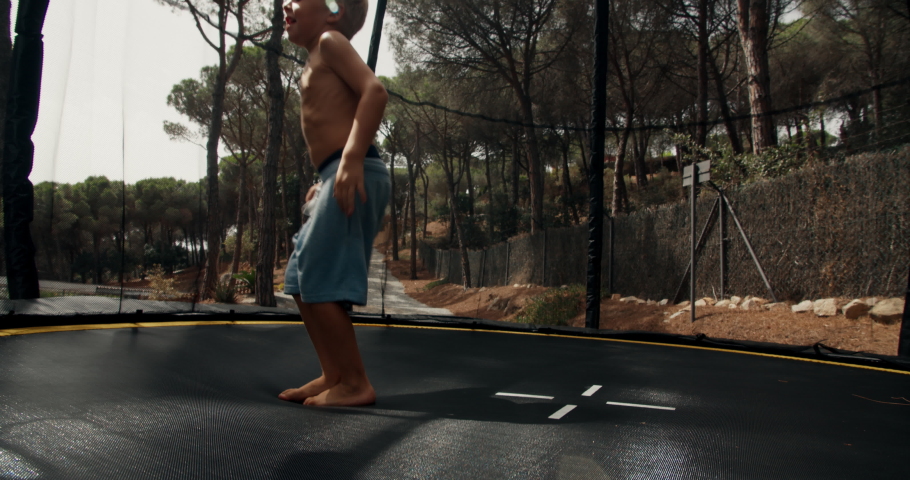 Little 4 years blonde boy is having fun during his summer vacations jumping on trampoline in forest of rural house terrain and smiling. Catalonia, Spain