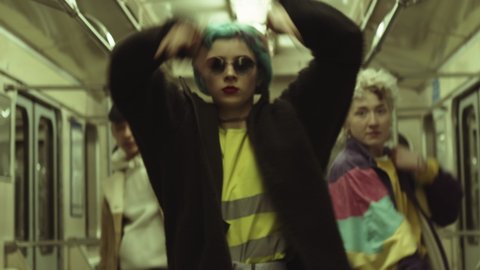 Handheld camera shot of young girl in trendy outfit and sunglasses dancing to camera with team in subway train