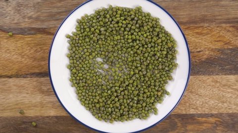 Green dry raw mung beans fall in a slow motion into a heap on a plate. Uncooked legumes. Close-up. Vigna radiata on a wooden table. Top view