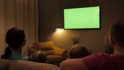 Rear View Of Family With Children Sitting On Sofa In Living Room Evening Watching Green Mock-up Screen TV Together. Family Sitting Together Sofa In Their Living Room Night Watching TV Green Screen. 