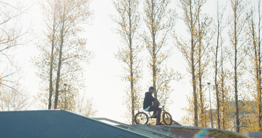 A young student smiles as he sits with bike on a ramp and a helmet on head at a skatepark, waiting for classmates with whom he can ride bikes and do stunts