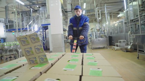 Industrial Dairy Factory Worker Moves Packaged Milk Cartons Production To Storage Facility. Worker With Production Packaged In Boxes On Pallet. Packaged Production Moved By Worker At Milk Plant