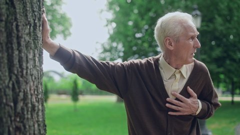 Elderly man feeling chest pain, leaning on tree and looking around, needs help