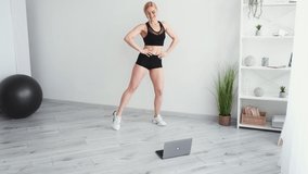 Online aerobic. Athletic woman. Home training. Lockdown sport. Happy sportive lady in black sportswear doing fitness exercise legs hands with laptop on floor in light room interior copy space.