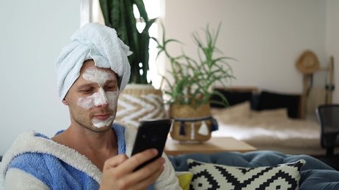 Young bearded man applying charcoal clay facial mask and looking at phone. Male skincare beauty treatment.