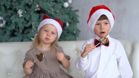 4k video portrait of two cute little children eating Christmas chocolate sweets happily. Craft chocolate handmade sweets in hands of kids. Brother and sister or friends wearing red holiday Santa hats