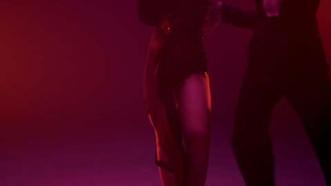 Unknown sensual woman lifting leg on dance partner indoors. Unrecognizable ballroom couple embracing inside. Passionate man and woman dancing in red light background. 