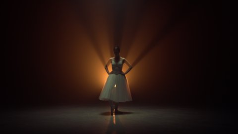 Young ballerina dancing on tiptoe with raised hands on dark stage. Flexible ballet dancer performing on spotlights background. Pretty woman wearing tutu skirt. Professional performer on theatre stage