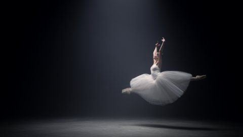 Gorgeous female dancer soaring in ballet dress on dark stage. Beautiful ballerina dancing in pointe shoes in spotlight. Young woman jumping on theatre stage classical dance. Elegance grace concept
