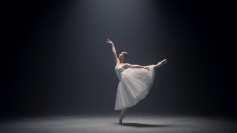 Graceful ballet dancer jumping on stage. Professional ballerina dancing on tiptoe in pointe shoes indoors. Sensual woman performer performing classical dance in tutu skirt in spotlight