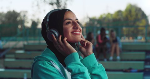 Cinematic shot of young arabic woman is having fun to listening to the music with earphones and dancing on basketball outdoor court after finishing friendly game match at sunset.