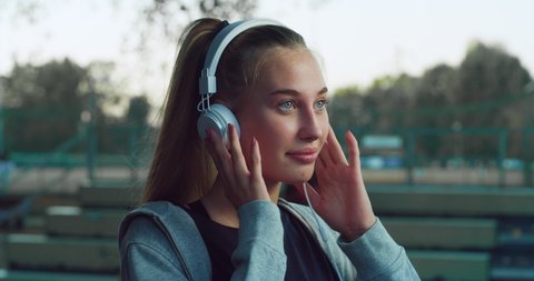 Cinematic shot of young blond hair woman is having fun to listening to the music with earphones and dancing on basketball outdoor court after finishing friendly game match at sunset.