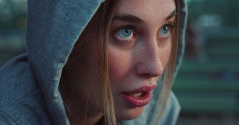 Cinematic close up shot of young blond hair woman with hood is taking break on basketball outdoor court after finishing friendly game match at sunset.