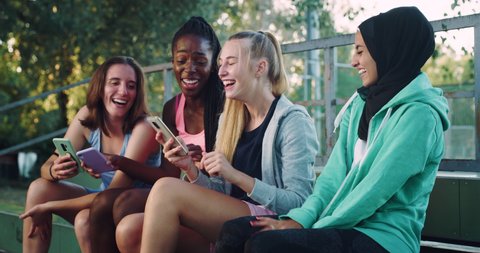 Cinematic shot of young happy girl friends of different ethnicities having fun to use smartphones together on basketball outdoor court after finishing friendly game match.