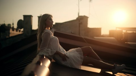 Portrait of gorgeous girl relaxing on rooftop. Side View of Young Attractive Blonde Woman in a white Dress Sitting on a Roof at Sunset, Wind Shuffles Her Beautiful Long Hair. Urban Cityscape View