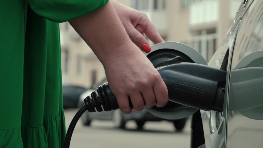 Unrecognizable woman in green dress replenishes energy of PHEV vehicle. Female hands opens charging port lid an electric car and plugging in power supply cable to charge EV | Shutterstock HD Video #1078797254