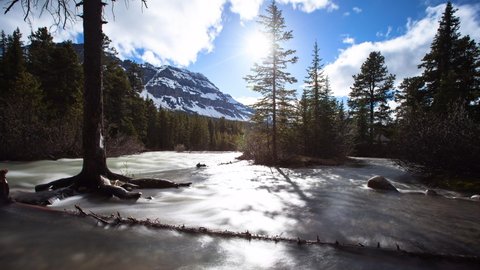 Time Lapse Shot Of Lake Flowing Amidst Forest At Banff National Park By Mountain Against Cloudy Sky - Alberta, Canada