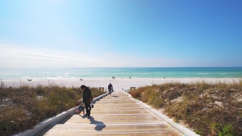 Seaside, USA - January 14, 2021: Point of view pov handheld walking in town city of Seaside, Florida gulf coast beach with wooden boardwalk steps and white sand with colorful emerald water