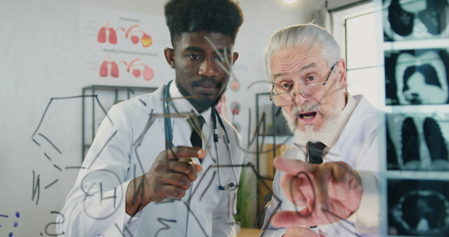 Attractive confident young african american medic-student attantive listening the explanation of chemical formula depicted on glass wall from respected skilled bearded professor in clinic classroom