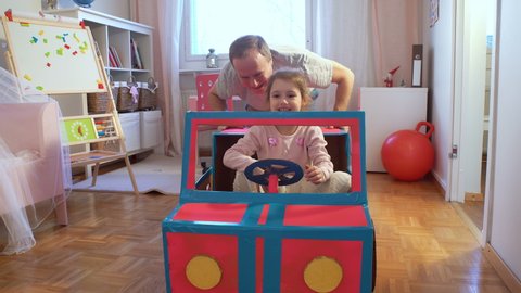 Funny little girl and her father are driving handmade cardboard car. Father and daughter playing in living room together.