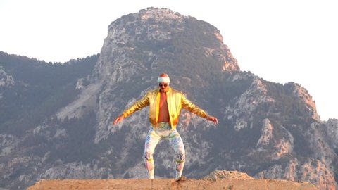 Young bald slender freak man caucasian in tight leggings, gold jacket, bandage on his head, dancing on roof at sunset, runs, has fun, behaves unusually. Slow motion video. Fun, humor, unusual people.
