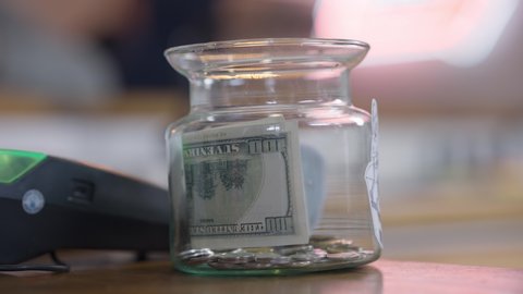 Close-up tips jar with female Caucasian hand putting cash inside. Unrecognizable cafe visitor leaving money for tips in cafeteria indoors